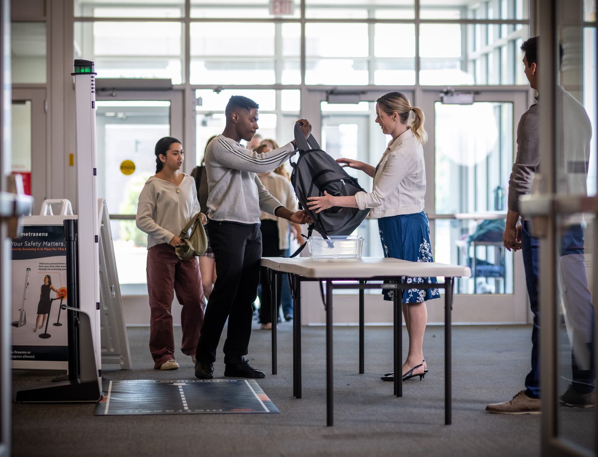 Students passing through a Metrasens Ultra screening lane at a school entrance while staff checks a backpack.